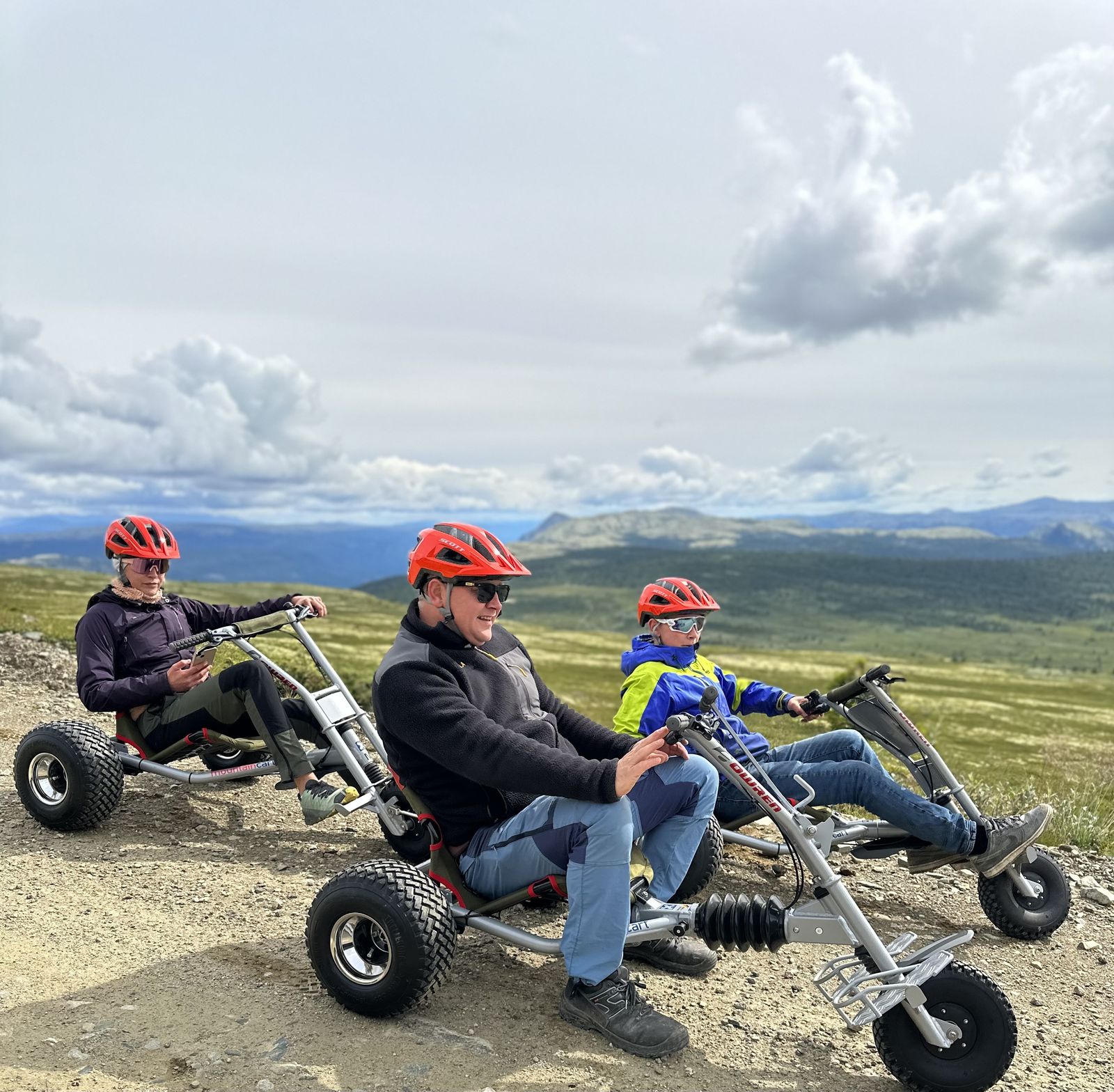 Voksen og to barn på mountaincarts med fjellpanorama bak